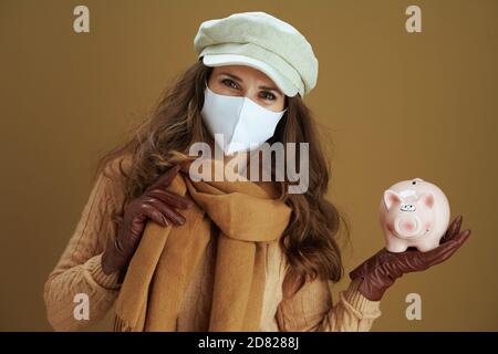 Hallo Herbst. Portrait der lächelnden modernen Frau in Schal mit Sparschwein und weiße medizinische Maske auf Bronze Hintergrund. Stockfoto