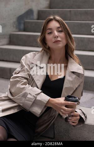 Attraktive schlanke junge Brünette in modischen Graben und schwarzen Kleid sitzt auf grauen Betontreppe mit einem braunen Papiertasse Kaffee. Stockfoto