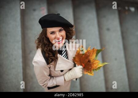 Hallo september. Obere Ansicht der lächelnden trendigen mittleren Alters Frau in beigem Trenchcoat und schwarzer Baskenmütze mit Herbst gelben Blättern im Freien in der Stadt in Stockfoto