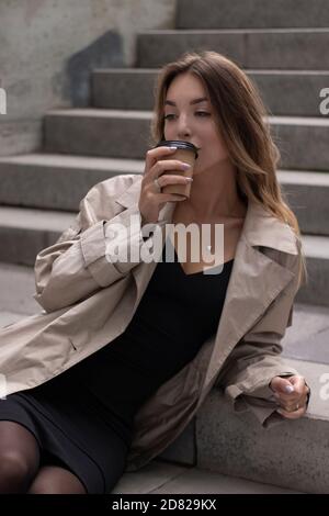 Attraktive schlanke junge Brünette in modischen Graben und schwarzen Kleid sitzt auf grauen Betontreppe mit einem braunen Papiertasse Kaffee. Stockfoto