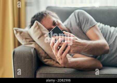 Deprimierter Mann mit Handy, während er auf dem Sofa im Wohnzimmer lag, Erwachsener trauriger einsamer Mann, der zu Hause schmollte Stockfoto