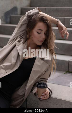 Attraktive schlanke junge Brünette in modischen Graben und schwarzen Kleid sitzt auf grauen Betontreppe mit einem braunen Papiertasse Kaffee. Stockfoto
