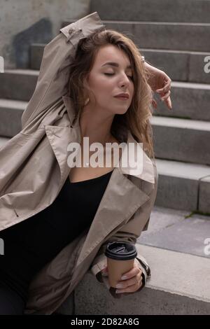 Attraktive schlanke junge Brünette in modischen Graben und schwarzen Kleid sitzt auf grauen Betontreppe mit einem braunen Papiertasse Kaffee. Stockfoto