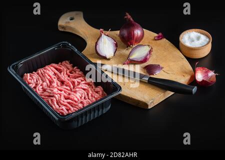 Hackfleisch ist ein Halbzeug in gewerblichen Verpackungen. Ein Messer und rote Zwiebeln liegen auf einem hölzernen Schneidbrett. Salzstreuer mit Salz. Schwarz-Bac Stockfoto