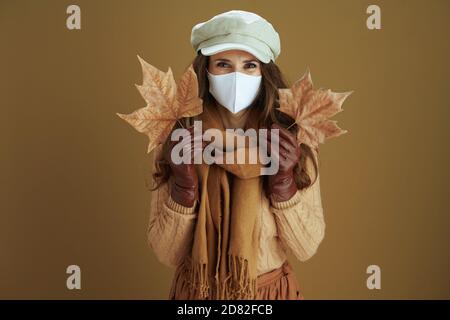 Leben während einer Coronavirus-Pandemie. Portrait von lächelnd elegante 40 Jahre alte Frau in Pullover mit weißen medizinischen Maske und gelben Herbst Ahornblätter iso Stockfoto