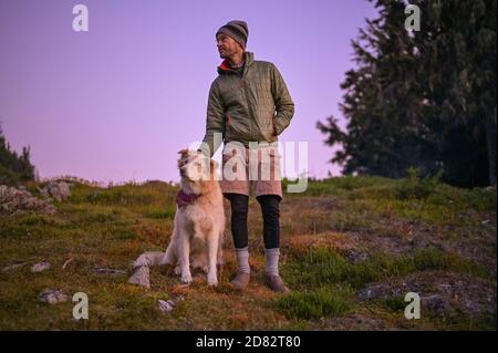 Wanderer im geschwollenen Mantel kratzen Hundeohr bei Sonnenuntergang Stockfoto