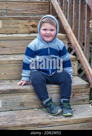 Ein glücklicher kleiner Junge sitzt auf Holztreppen und trägt einen kapuzenjacke und ein großes Lächeln Stockfoto