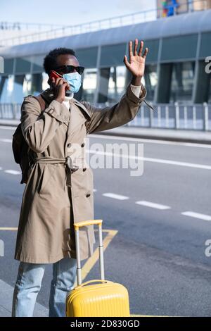 Afro-amerikanischer Reisender Mann mit Koffer tragen Gesichtsmaske steht im Flughafen-Terminal, im Gespräch auf Handy, rufen Taxi, heben die Hand. Warten auf c Stockfoto