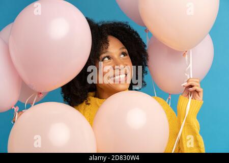 Nahaufnahme von glücklichen dunkelhäutigen Frau unter pastellrosa Luftballons mit Freude und Glück, feiert Geburtstag, schauen, trägt gelben Pullover, Stan Stockfoto