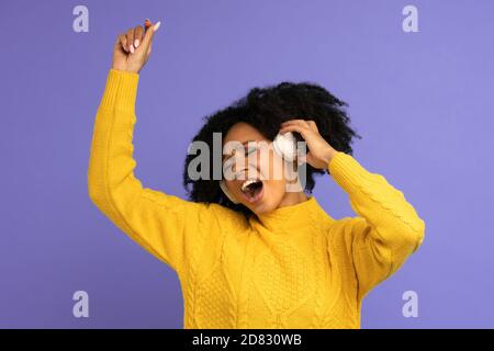 Energiegeladene Birazialfrau singt mit, lächelt breit, trägt kabellose Kopfhörer, hat gute Laune, isoliert auf violettem Hintergrund. Glücklich dunkelhäutig gelockt Stockfoto