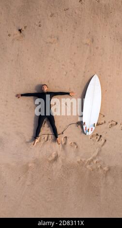 Blick von oben auf einen Mann Surfer in Neoprenanzug mit einem Surfbrett auf dem Sand am Strand liegen. Müde nach dem Surfen. Sich ausruhen Stockfoto