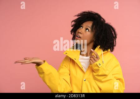 Biracial lockige Frau in gelben Regenmantel hat gute Laune, sieht aus, wenn es draußen regen, Blick auf leeren Raum für Werbung, Werbe-Inhalte, ist Stockfoto