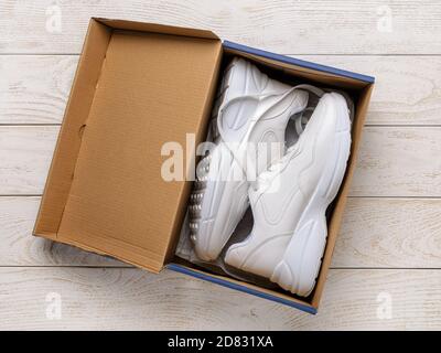 Zwei weiße Turnschuhe mit grober Sohle in einer braunen Kartonschachtel auf dem weißen Holzboden. Offene Box mit neuen bequemen Schuhen für aktiven Lifestyle, Fitness Stockfoto