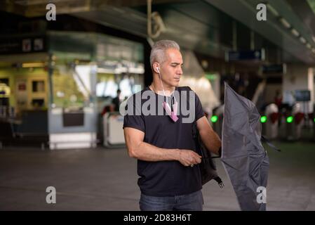 Schöner persischer Mann mit grauen Haaren öffnen Regenschirm an der Skytrain-Station Stockfoto