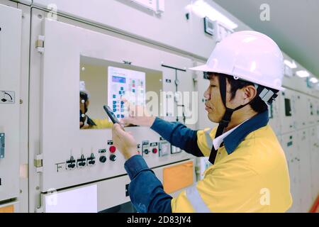 Ingenieur mit gelber Uniform und weißem Schutzhelm, der elektrische Systeme in einem großen Kraftwerk inspiziert. Stockfoto