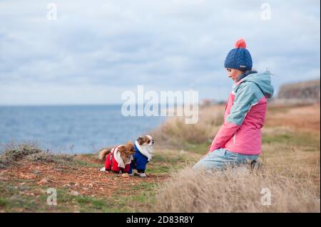 Nachdenkliche gesunde Sport junge Frau in Hut und Track Suit Sitzen vor zwei gekleideten kleinen Haustieren chihuahua Hunde auf Kälte Wetter Frühling Meer Küste Stockfoto