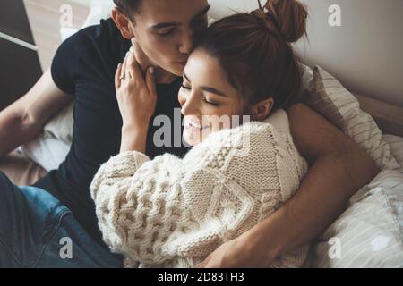 Kaukasischer Mann küsst seine nette Frau in einem weißen gekleidet Strickpullover umarmt im Bett Stockfoto