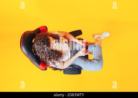 Frau, die hinter einem Spielstuhl in ihren Händen sitzt und roten Gamepad auf gelbem Hintergrund hält, Blick von oben Stockfoto