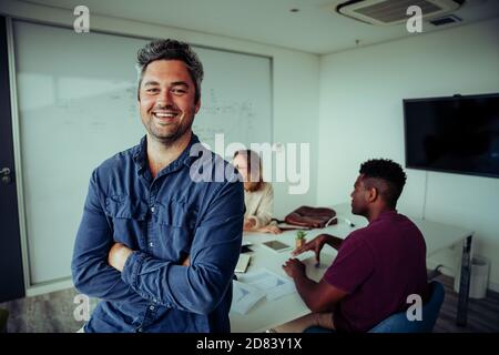 Kaukasischer Geschäftsmann lächelt und steht selbstbewusst mit den Querarmen davor Von hart arbeitenden Kollegen Stockfoto