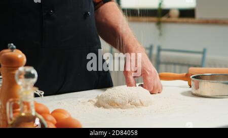 Baker Zugabe von Mehl auf den Haufen von Hand Zubereitung Brotteig. Nahaufnahme eines älteren Küchenchefs im Ruhestand mit bonete und einheitlicher Streuselung, Sieben von überzogenen Rohzutaten beim Backen von hausgemachter Pizza und Hackbäcken. Stockfoto