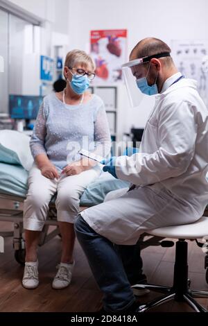 Arzt liest Diagnose der älteren Frau Krankheit trägt Gesichtsmaske gegen covid Pandemie als Sicherheitsmaßnahme im Krankenhaus. Medizinische Arztkonsultation während der globalen COVID-19-Krise. Stockfoto