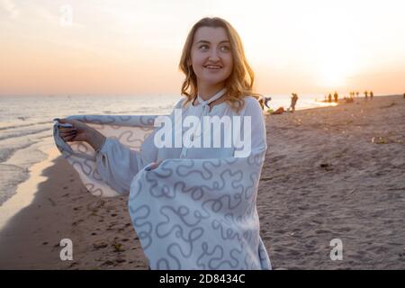 Junge schöne Frau in eine warme Decke eingewickelt, warm und gemütlich am Strand. Am Strand bei Sonnenuntergang laufen, sich frei und glücklich fühlen. Stockfoto