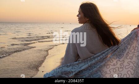 Junge schöne Frau in eine warme Decke eingewickelt, warm und gemütlich am Strand. Am Strand bei Sonnenuntergang laufen, sich frei und glücklich fühlen. Stockfoto