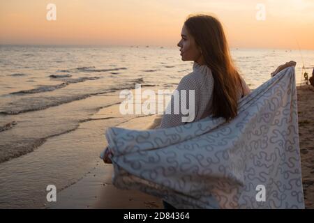 Junge schöne Frau in eine warme Decke eingewickelt, warm und gemütlich am Strand. Am Strand bei Sonnenuntergang laufen, sich frei und glücklich fühlen. Stockfoto