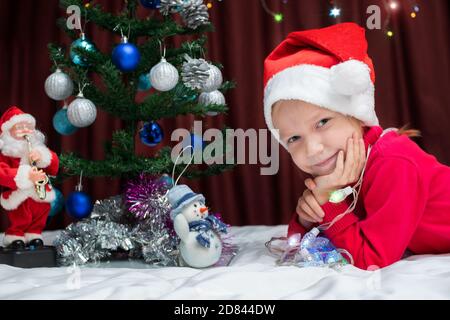 Ein süßes kaukasisches blondes Mädchen in einem roten Pullover und Weihnachten sieht Sie verträumt an, das Mädchen liegt neben einem verzierten Weihnachtsbaum, einem Spielzeug Schneemann und Stockfoto