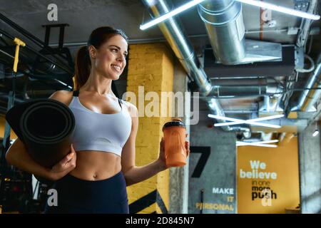 Porträt einer jungen schönen Fitness-Frau in Sportswear hält Yoga-Matte und eine Flasche Wasser, Blick auf das Fenster beim Training im Fitnessstudio. Sport, Wellness und gesunder Lebensstil Stockfoto