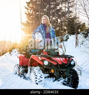 Portrait von schönen blonden Mädchen, trägt blaue Winterjacke, hält ihren Helm unter der Achsel, stand auf roten Offroad-Vierrad ATV bei Sonnenuntergang. Konzept der aktiven Freizeit und Winteraktivitäten. Stockfoto