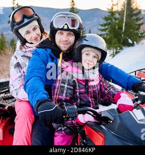 Fröhliche Familie in Helmen lächelt vor der Kamera, während auf All-Terrain-Fahrzeug mit entzückenden Tochter sitzen. Nettes Mädchen genießen Quad Radfahren mit Eltern. Konzept der aktiven Freizeit und Winteraktivitäten. Stockfoto