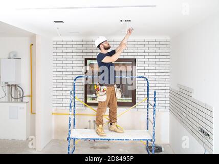 Elektriker Baumeister bei der Arbeit, Installation der Lampen in der Höhe. Professionell in Overalls mit einem elektrischen Werkzeug. Auf dem Hintergrund der Reparaturstelle. Stockfoto