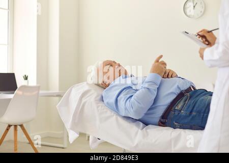 Älterer Mann, der im Krankenhausbüro auf dem Bett liegt und mit ihm spricht Arzt, der die Gesundheitskarte ausfüllt Stockfoto