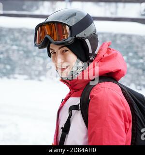 Seitenansicht der charmanten jungen Frau in Winterjacke Blick auf die Kamera und lächelnd. Fröhliche Dame mit Rucksack trägt Schutzhelm und Skibrille. Konzept von Skifahren und Wintersport. Stockfoto