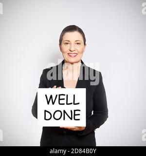 Lächelnde weibliche Chefin in schwarzem Business-Outfit hält eine weiße Karte mit Text GUT GEMACHT. Stockfoto