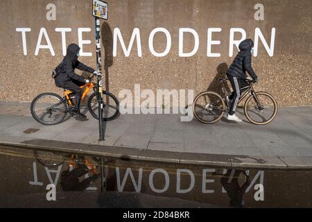 Zwei junge schwarze Männer fahren am 26. Oktober 2020 in London, England, mit dem Fahrrad an der Brütalistischen Betonarchitektur der Tate Modern Art Gallery auf der Southbank vorbei. Stockfoto