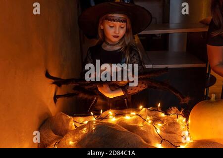 Kind Mädchen Vorbereitung Halloween in Hexenkostüm zu feiern. Große Spinne in der Hand als Dekor. Kürbis beleuchtet und Girlande auf Tisch. Stockfoto