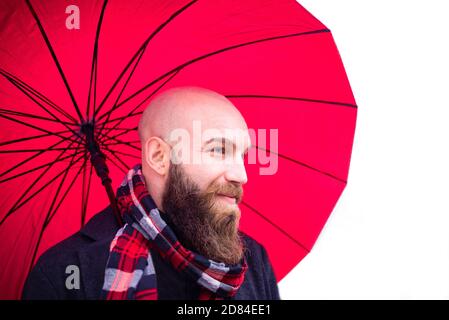 Bolt bärtiger Mann trägt einen Schal und warme Winterkleidung Halten Sie einen roten Schirm als Hintergrund.lächelnde junge Erwachsene auf Ein weißes Szenario in einem Studio Stockfoto