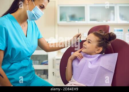 Kinderzahnarzt. Kleine, neugierige Mädchen sitzt im Stuhl in der Zahnklinik und im Gespräch mit freundlichen Arzt in medizinische Maske Stockfoto