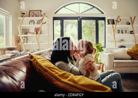 Verspielte junge Frau küssen Kleinkind Mädchen, während auf dem Sofa sitzen Zu Hause Stockfoto