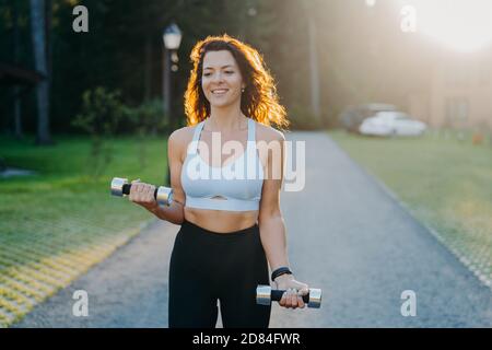 Foto von schlanken Brünette junge Frau hebt Hanteln hat Morgen Workout posiert gegen den Sonnenaufgang, gekleidet in 3/4-Oberteil und Leggings Funktioniert auf Arme Muskel Stockfoto