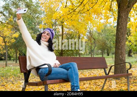 Junge Frau mit lockigen schwarzen Haaren und warm in Pullover Und Wollhut, die ein Selbstporträt mit ihrem Handy Während Sie lächeln, sitzen auf einer Holzbank in einem Stockfoto