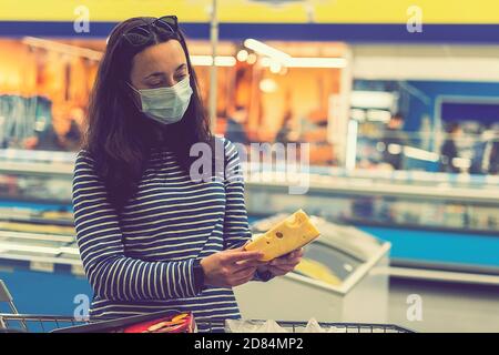 Frau in schützender medizinischer Maske in einem Supermarkt wählt Käse. Pandemie und Covid-19-Konzept. Hand des Käufers mit einem Stück Käse im Laden Stockfoto