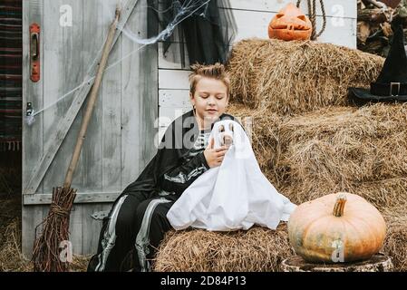 Ein Junge in einem Skelett Kostüm mit einem Hund in Ein Geisterkostüm auf der Veranda eines Hauses dekoriert Um eine Halloween Party zu feiern Stockfoto