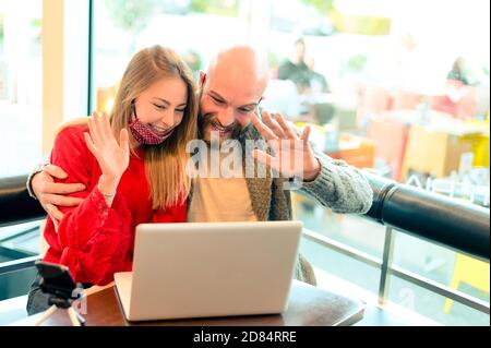 Portrait von Studenten mit Gesichtsmaske mit Laptop für ein Online-Meeting in Video-Chat in einem Café in Coronavirus Zeit - Business-Team winkt Hand an vide Stockfoto