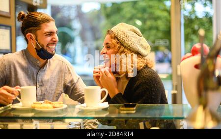Schöne glücklich liebende Paar mit Gesichtsmaske genießen Frühstück in Ein Café in der Coronavirus-Zeit -zwei lächelnde Freunde trinken Capucino und essen Cookie A Stockfoto