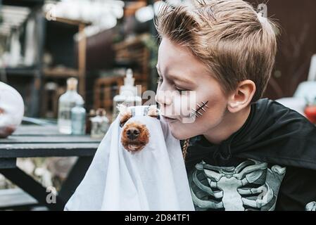 Ein Junge in einem Skelett Kostüm mit einem Hund in Ein Geisterkostüm auf der Veranda eines Hauses dekoriert Um eine Halloween Party zu feiern Stockfoto