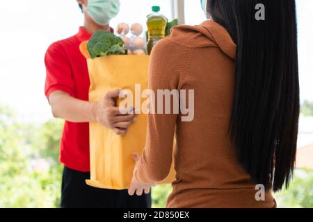 Asiatische Lieferung Mann aus dem Supermarkt trägt eine Gesichtsmaske und hält eine Tasche mit frischen Lebensmitteln, Gemüse und Obst für die Kunden zu Hause geben. Conce Stockfoto