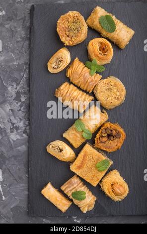 Traditionelle arabische Süßigkeiten (Kunafa, Baklava) auf schwarzem Schiefertafel auf schwarzem Betongrund. Draufsicht, flach liegend, Nahaufnahme. Stockfoto